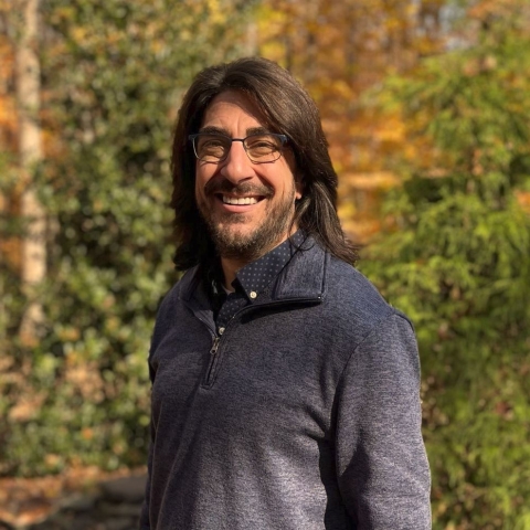 A person standing against a background of trees wearing glasses, and smiling.