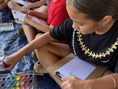Three children sitting down, painting waterbird art with paint brushes and watercolors.