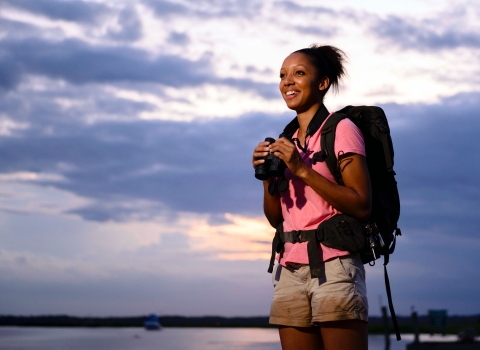 A young Black woman wearing a backpack and holding binoculars and smiling, looks off into the distance under a cloudy blue sky.