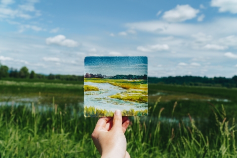 A hand holding up a tiny watercolor of the scene behind it