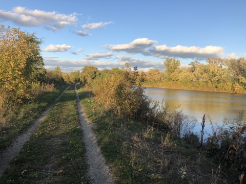 The hogback ridge trail next to a pond stretches out into the misty evening light. Lush vegetation is set infront of a blue sky with clouds.