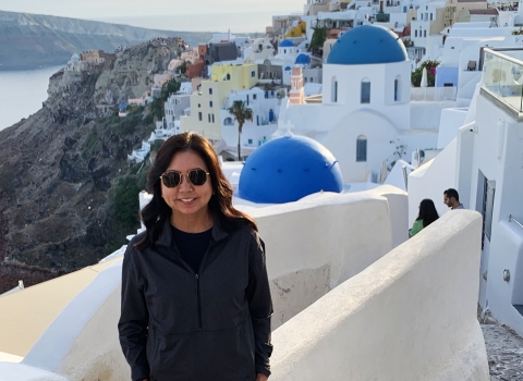 Susan Machida stands along cobblestone steps. The buildings of Santorini, Greece, adorn the background. Their ivory white colors contrast against the brown cliffsides. 