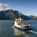 Blue and white ship sails towards land over calm blue waters and a blue sky with a few clouds.