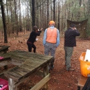 A man and a women holding firearms face the woods with a man with a bright orange vest on in between them.