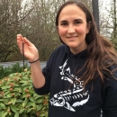 Julie Harris, Biometrician at CRFWCO, holding a juvenile Pacific Lamprey in the palm of her hand while wearing a black hoodie. Julie is standing next to bushes in a natural setting