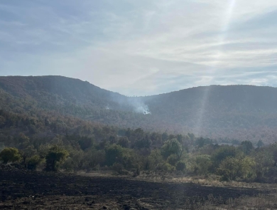 An arid lansdcape of hills covered with juniper and burn scars, with a large smoke from a wildfire visible in the center