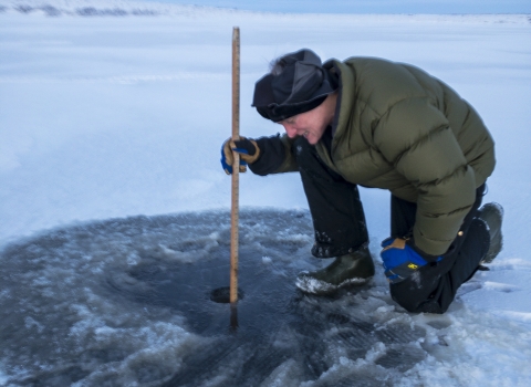 a man crouches near a small round hole in the ice and uses a yardstick to measure
