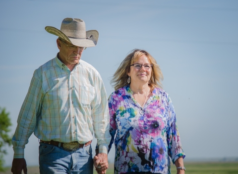 A man wearing a cowboy hat and a woman walk hand-in-hand across a grassy field