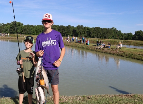 2 Youths, one holding catfish stringer and the other holding a fishing rod and reel in front of pond.