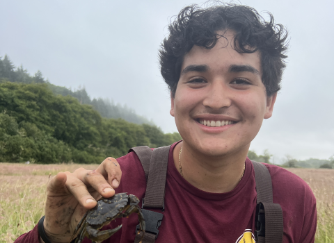 AmeriCorps service member in waders holding a European green crab. Marsh vegetation, trees, and overcast sky in the background.