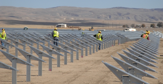 rows of gray solar panels stand out against the dry desert background. Solar panel installers wearing yellow vests work on the panels.