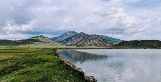 A lake abuts green grass, in the background are small mountains
