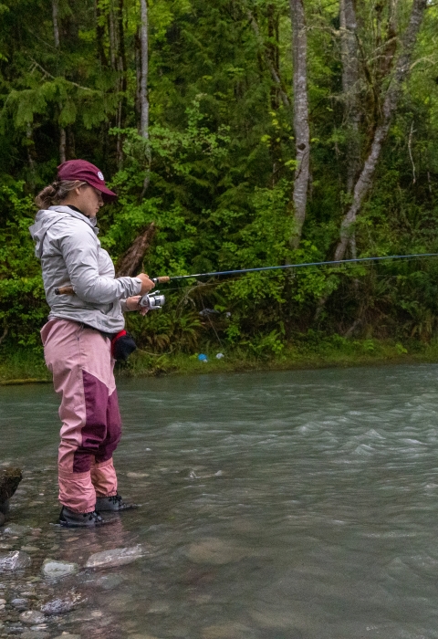 Fisheries intern, Hannah Ferwerda, wading in water and reeling a fishing rod as she samples river for fish.