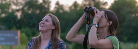 Two women enjoy birdwatching at the National Conservation Training Center in West Virginia. One looks up at trees through binoculars.