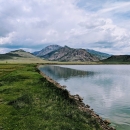 A lake abuts green grass, in the background are small mountains