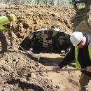 Two people in hard hats and vests shovel dirt in front of a metal culvert.