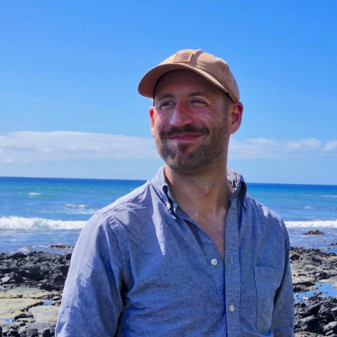 A man in a blue shirt and tan hat stands in front of the ocean