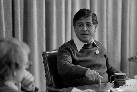 Black and white picture of a man with dark hair and eyes sitting at a meeting table. He is gesturing and speaking to another person in the foreground. He wears a sweater over a collared shirt. There is a button attached to the sweater over his left breast. 