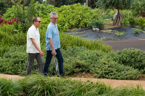 Two older men walk down a path surrounded by lush greenery 