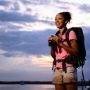 A young Black woman wearing a backpack and holding binoculars and smiling, looks off into the distance under a cloudy blue sky.