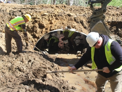 Two people in hard hats and vests shovel dirt in front of a metal culvert.