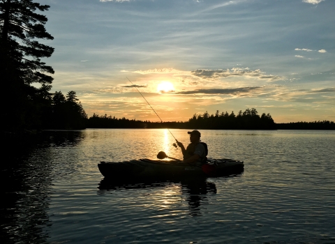 a sillhouette of a person kayak fishing while floating on a large body of water. The sun sets behind them and the sky is colored blue and yellow