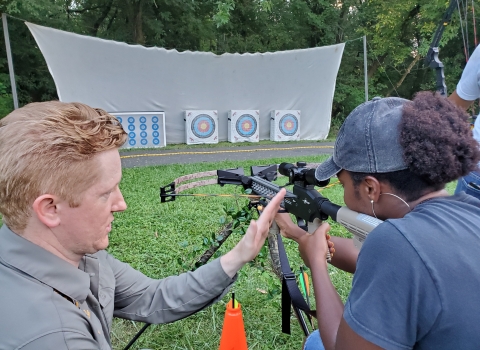 A man teaching a woman how to handle a crossbow while she aims at an archery target