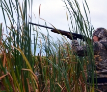 Female standing in tall grass wearing camouflage hunting outfit holding a rifle and aiming to the left.