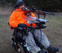 A hunter dressed in orange enjoys the outdoors with the aid of a wheelchair and mounted gun.