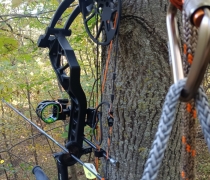 An archery bow sits on a hunting tree stand facing green vegetation with a carabiner in the foreground.