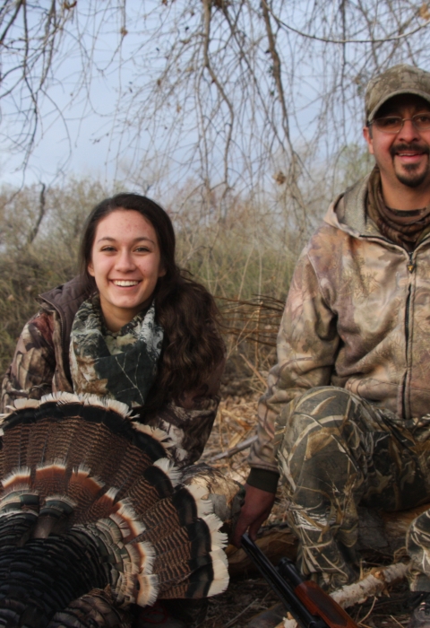 A young woman and a man in camouflage sit on a ground in a wooded area with a wild turkey