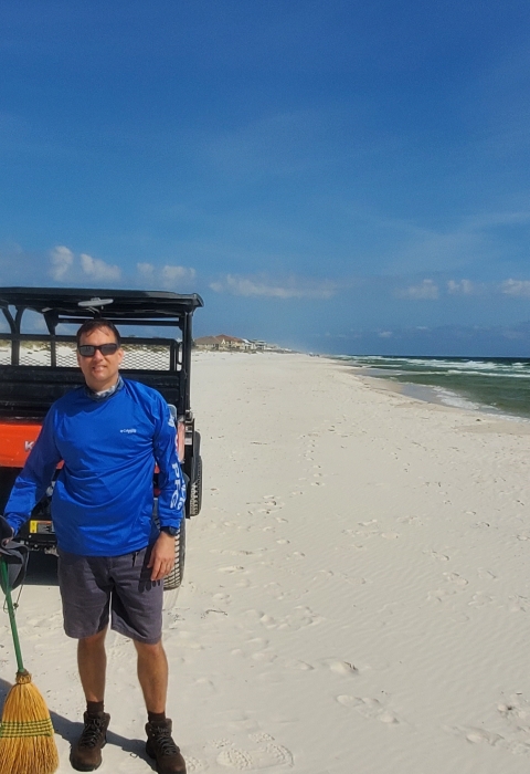 Under a partly sunny sky, facing the camera is a man wearing a blue shirt standing on a white sand beach in front of an off road beach club car.