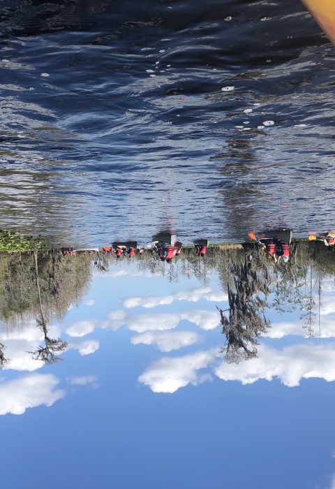 The bow of a kayak floats in the corner of a photo. Multiple canoes are seen in the background, filled with happy students paddling on the Okefenokee Swamp. 