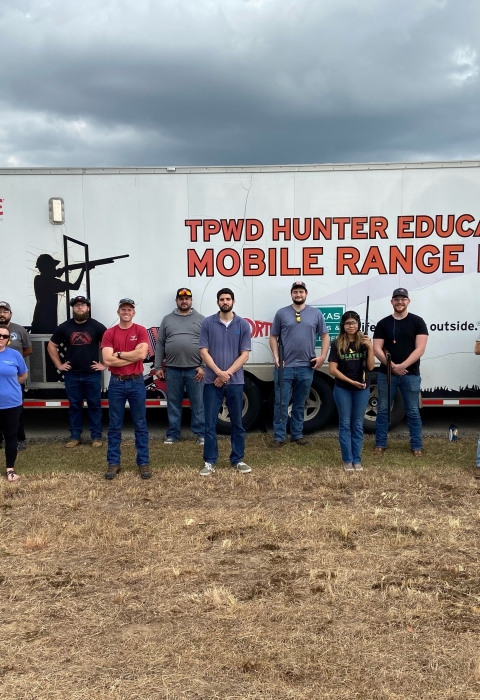Shooting sport youth participants and mentors stand in front of the white Hunter Education Mobile Range Trailer