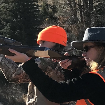 Two women in hunting gear side by side holding long guns side in shooting position