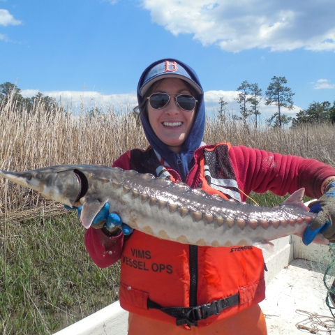 A smiling woman wearing an orange life vest, sunglasses and hat holds up a large fish