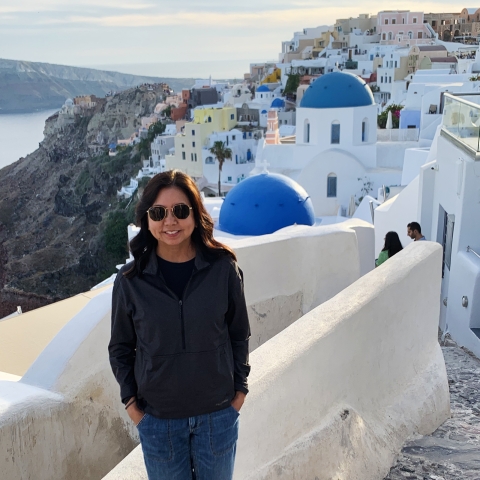 Susan Machida stands along cobblestone steps. The buildings of Santorini, Greece, outline the background. 