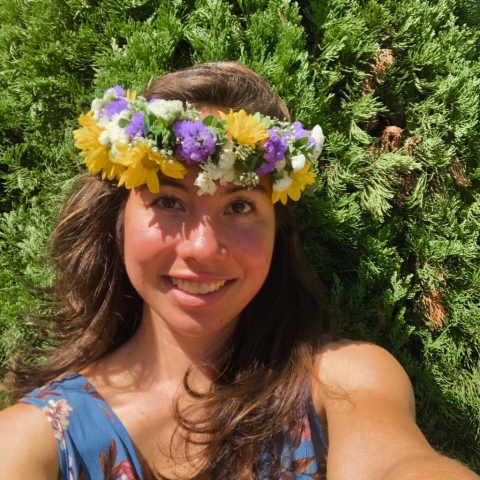 A person with long brown hear wearing a traditional lei on their head takes a selfie.