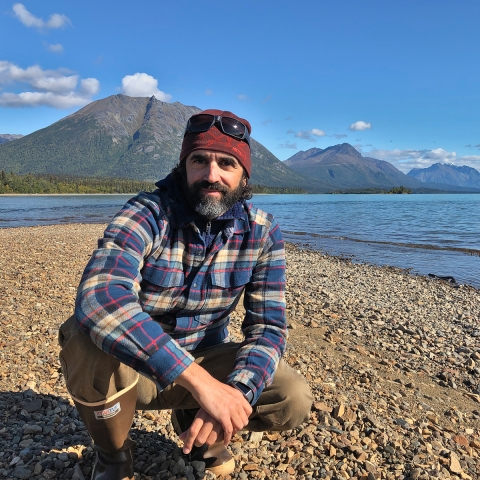 A person crouches on a gravel beach on the shore of a turquoise lake with mountains on the horizon the distance.