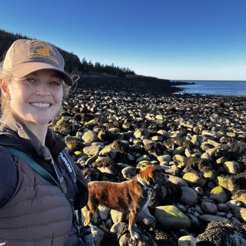 woman standing on large rocks by shore with dog by side