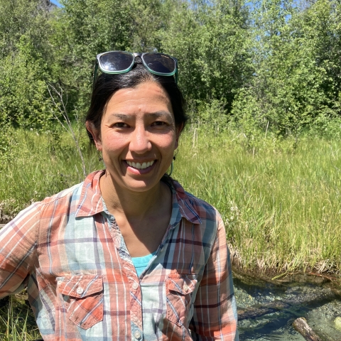 woman in an orange and while plaid shirt stands in front of a grassy field with trees