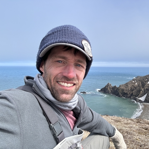 A person smiles while taking a selfie from a cliff overlooking an ocean coastline with jagged snow-covered cliffs.