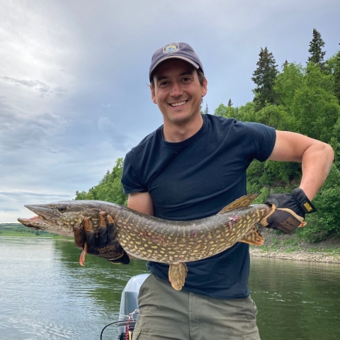 A person holds aa long fish with both hands in front of their torso with a body of water and vegetated hill behind them. They smile.