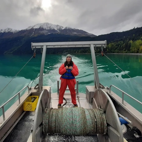 A person standing on a boat wearing bright orange with bright green water and mountains in the background