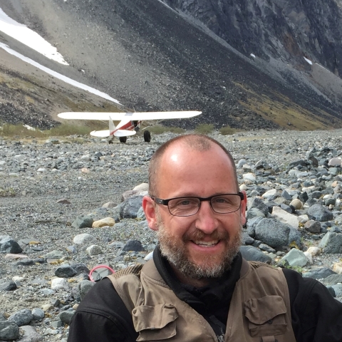 A close-up of a man with a rocky mountain slope and small plane in the background