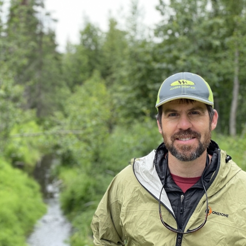 man in a hat standing by a wooded stream