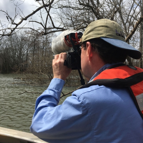 Man in blue shirt and life vest with ball cap on backwards shooting video while riding in boat