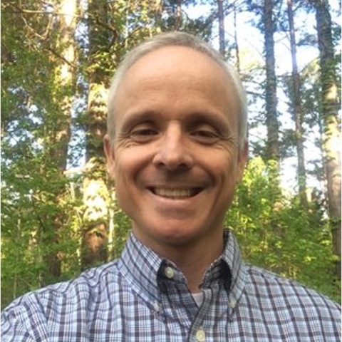 Keith smiles in a blue plaid button down in a forested area