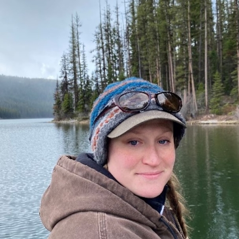 Kiandra smiling with a blue knit cat, sunglasses, and a brown hoodie. She stands infront of a body of water and trees. 