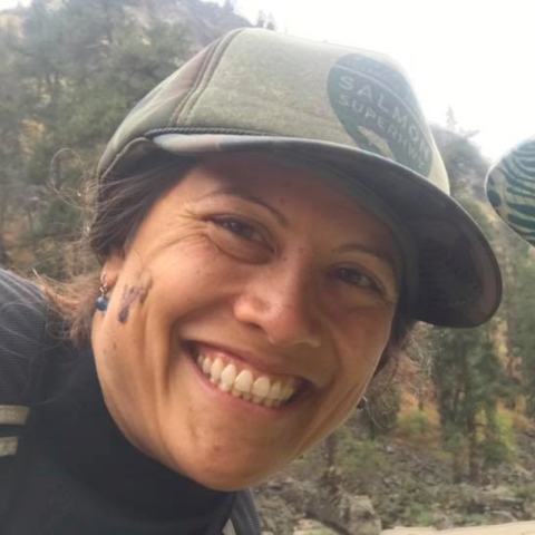 A woman wearing a hat and smiling with mountains and trees in the background